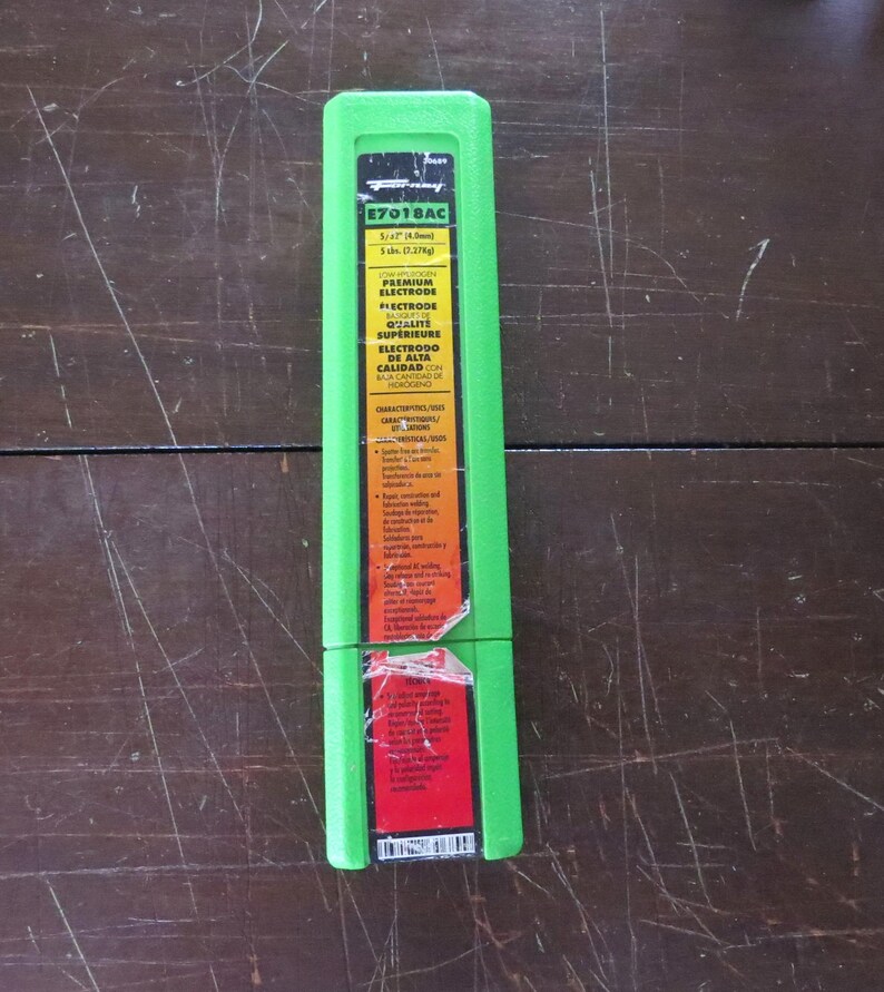 May include: A green rectangular package of E7018AC premium electrodes, 5 lbs (2.27kg). The package has a yellow and red label with text in English and Spanish. The package is on a dark brown wooden surface.