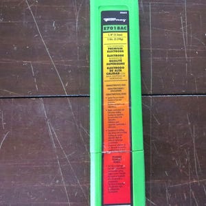 Puede incluir: Un paquete verde de electrodos premium de bajo hidr&oacute;geno E7018AC. El paquete es rectangular con un marco verde y una etiqueta roja y blanca. La etiqueta incluye texto sobre las especificaciones del electrodo, incluyendo 3,2 mm y 2,27 kg.