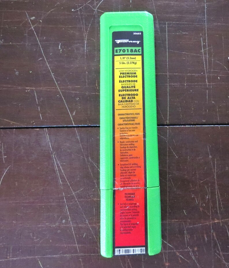 Puede incluir: Un paquete verde de electrodos premium de bajo hidr&oacute;geno E7018AC. El paquete es rectangular con un marco verde y una etiqueta roja y blanca. La etiqueta incluye texto sobre las especificaciones del electrodo, incluyendo 3,2 mm y 2,27 kg.