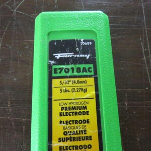 May include: A green plastic container with a black and yellow label. The label features the "Forney" logo, product information, and the text "E7018AC". It also indicates the electrode size as 4.0mm and weight as 2.27Kg.
