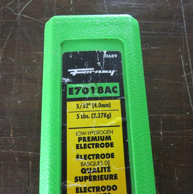 May include: A green plastic container with a black and yellow label. The label features the "Forney" logo, product information, and the text "E7018AC". It also indicates the electrode size as 4.0mm and weight as 2.27Kg.