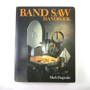 May include: A book titled "BAND SAW HANDBOOK" with a black cover and orange lettering. The cover features an illustration of a band saw with a wooden workpiece. The author's name, Mark Duginske, is printed at the bottom.