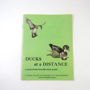 May include: A light green softcover book titled DUCKS at a DISTANCE a waterfowl identification guide. The cover features a black and white illustration of a duck in flight and a colorful illustration of a duck swimming on the water.