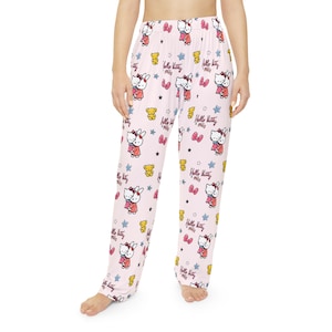 Op de afbeelding: Lichtroze pyjamabroek met een herhaald patroon van Hello Kitty-personages, strikken, sterren en teddyberen. De tekst "Hello Kitty" staat ook op de broek. De pyjamabroek is gemaakt van een zacht materiaal.