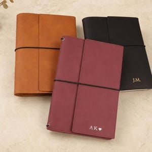 Peut inclure: Trois carnets rectangulaires avec des couvertures enveloppantes en brun clair, bordeaux et noir. Chaque carnet est maintenu par un élastique noir. Le carnet bordeaux porte l'inscription AK avec un cœur, et le carnet noir porte l'inscription J.M.