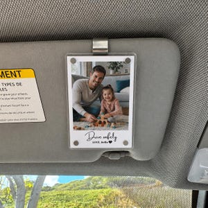 Könnte beinhalten: Ein rechteckiger, durchsichtiger Acryl-Fotorahmen mit einem Metallclip, der an einer Sonnenblende im Auto befestigt ist. Der Rahmen zeigt ein Foto eines Erwachsenen und eines Kindes beim Spielen mit Holzklötzen sowie den Text Drive safely LOVE, Bella.