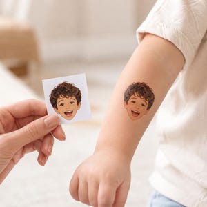 May include: A square white paper sheet featuring a colorful illustration of a smiling child with curly brown hair, alongside a matching temporary tattoo applied to an arm, depicting the same illustration of the child's face with a joyful expression.