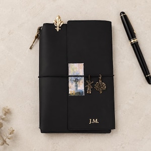 May include: A black leather journal featuring the initials J.M. embossed in gold, a gold fleur-de-lis clip, a postage stamp, and two metal charms shaped like a windmill and a tree attached to a black elastic band, accompanied by a black fountain pen.