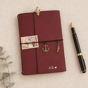 Peut inclure: Un journal rouge foncé avec un clip fleur-de-lis doré sur le dessus. Un élastique noir maintient un timbre rectangulaire, une breloque ancre dorée et une breloque Statue de la Liberté dorée. La couverture porte les lettres AK et un cœur.