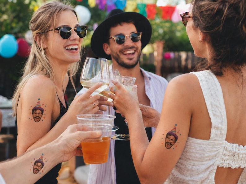 May include: Temporary tattoos featuring a cartoon face wearing a colorful party hat. The design includes a smiling person with glasses and a mustache, topped with a multi-colored cone hat, applied to the skin of three different individuals.