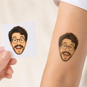 May include: A temporary tattoo featuring a cartoon-style illustration of a person with dark wavy hair, a mustache, and round glasses, shown with a wide, happy expression. The same illustration is displayed on a square white paper backing.