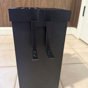 May include: A black rectangular storage container with a handle-like design on the front. The container has a grid-like top and is made of a matte material. The background features a wooden wall and a white door.
