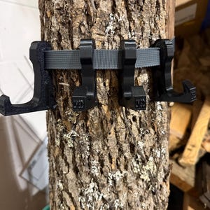 May include: A set of four black plastic hooks attached to a grey fabric strap, secured around a rough, textured piece of tree bark. Each hook features a distinct J-shape design with a flat base and a small upward-curving tip for hanging items.