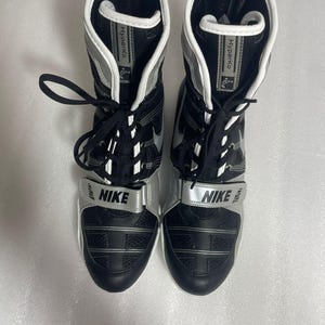 May include: Pair of black and silver Nike Hyperko boxing shoes. The shoes have a high-top design with a silver strap across the top of the foot that says "NIKE". The shoes have black laces and a white interior.