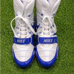 May include: Pair of white and blue Nike wrestling shoes. The shoes have a blue strap with the Nike logo, white laces, and blue accents. The shoes are on a green surface.