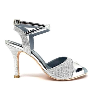 Op de afbeelding: Zilveren sandaal met hoge hak en glitterafwerking. Voorzien van een enkelbandje met gesp en een spitse neus. De hak is ongeveer 7.6 cm hoog.