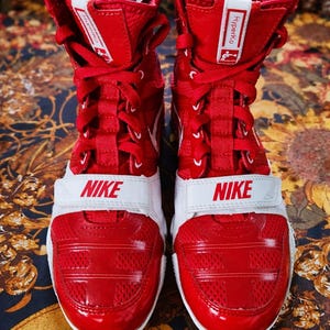 May include: Pair of red and white Nike boxing shoes. The high-top sneakers feature a red upper with a white strap across the midfoot, displaying the Nike logo. The shoes have red laces and a white tongue. The word "Hyperko" is printed on the side.