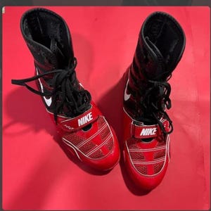 May include: Pair of red and black Nike wrestling shoes. The shoes have a high-top design with black laces, a white Nike swoosh, and a red strap with the Nike logo. The shoes are on a red background.