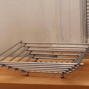 May include: A square, stainless steel wire rack with a grid pattern, elevated by small, spherical feet. The rack is designed with parallel bars and is suitable for cooling baked goods or serving appetizers. It sits on a wooden surface.