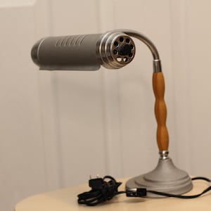 Puede incluir: Una lámpara de escritorio vintage con una pantalla y base de metal gris, un cuello de cisne plateado y un mango de madera. La lámpara tiene un cable de alimentación y un enchufe negros. La lámpara está sobre una superficie clara.