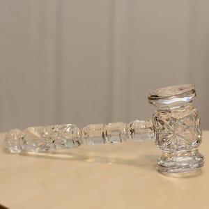 May include: A clear glass gavel with a faceted design, resting on a light-colored surface. The gavel's head and handle are intricately cut, reflecting light. The background is a neutral, blurred setting.
