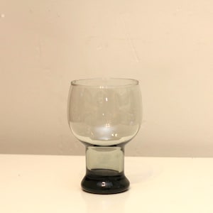 May include: A clear glass goblet with a dark gray base and stem. The bowl of the glass is wide and rounded, tapering slightly towards the rim. The glass sits on a white surface against a neutral background.