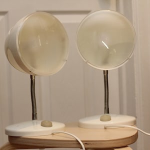 May include: Two vintage white desk lamps with flexible metal necks and round, frosted glass shades. The lamps are on a light-colored wooden surface, with a white cord extending from each base. The lamps are turned on.