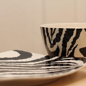 Op de afbeelding: Een keramische kom en vierkante bordenset met een zwart-wit zebra streepmotief. De kom is rond met een brede opening. Het bord is vierkant met afgeronde hoeken. De set staat op een lichtgekleurd oppervlak.