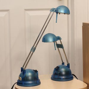 May include: Two blue desk lamps with adjustable arms and dome-shaped shades. The lamps have a metallic silver finish on the arms and a blue base. The lamps are on a light-colored surface.