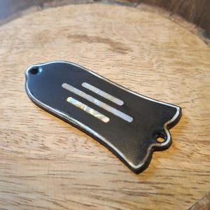 Peut inclure: Un cache truss rod de guitare noir en forme de cloche, orné de trois bandes horizontales incrustées en nacre blanche. La pièce présente deux petits trous de montage circulaires en haut et en bas ainsi qu'un fin contour blanc.