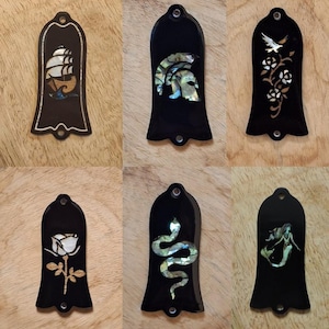 May include: Six black bell-shaped guitar truss rod covers featuring iridescent mother-of-pearl inlays. Designs include a sailing ship, a Spartan helmet, a bird with roses, a single rose, a coiled snake, and a mermaid, each with two mounting holes.