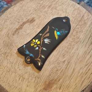 Puede incluir: Un colgante negro, alargado y con forma de campana, que presenta un diseño incrustado de una flor amarilla y un colibrí azul. La pieza tiene un acabado brillante e incluye dos pequeños orificios circulares en la parte superior e inferior.