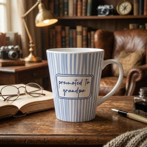 Peut inclure: Une tasse en céramique blanche avec des rayures verticales bleues et le texte "promoted to grandpa" dans un cadre rectangulaire. La tasse est sur une table en bois avec un livre, des lunettes et un stylo.
