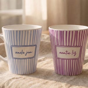 May include: Two ceramic mugs with a tapered shape and vertical stripes. The left mug is light blue with the text "uncle jim" in a rectangular frame. The right mug is purple with the text "auntie liz" in a matching rectangular frame.