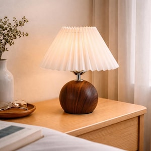 May include: A small table lamp with a white pleated lampshade and a round, dark wood base. The lamp sits on a light wood nightstand. A vase with greenery and a wooden tray with glasses are also visible.
