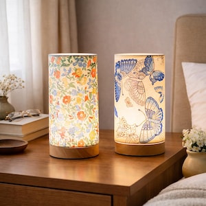 Puede incluir: Dos lámparas de mesa cilíndricas con bases de madera. Una lámpara presenta un estampado floral en naranja, amarillo y azul. La otra lámpara tiene un diseño de mariposas en azul y blanco. Ambas lámparas están iluminadas.