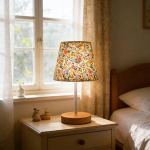 May include: A table lamp with a floral patterned lampshade. The lampshade features a colorful array of flowers on a cream background. The lamp has a white stem and a round wooden base. The lamp sits on a white nightstand.