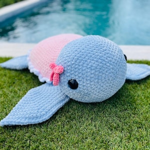 May include: A handmade, plush crochet turtle in shades of blue and pink. The turtle has a light blue head and flippers, a pink shell, and a small pink bow. The turtle is lying on green grass.