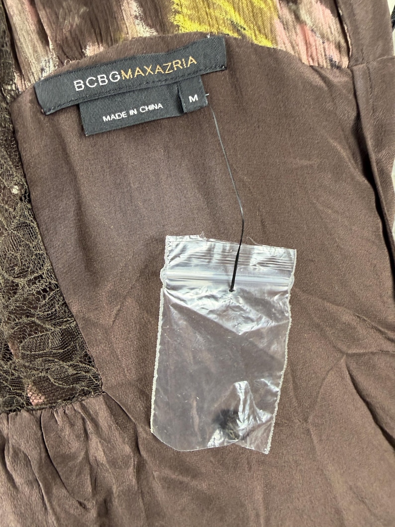 May include: Close-up of a brown dress with a BCBGMAXAZRIA label. The label is black with white text and the size is M. The dress has a lace detail and a clear plastic bag attached.