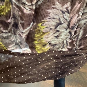 May include: Close-up of a brown fabric with a floral pattern in shades of green, pink, and gray. The bottom edge features a dark brown band with white polka dots. The fabric appears to be draped.