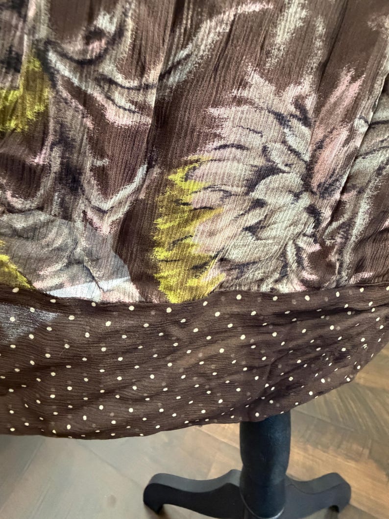 May include: Close-up of a brown fabric with a floral pattern in shades of green, pink, and gray. The bottom edge features a dark brown band with white polka dots. The fabric appears to be draped.