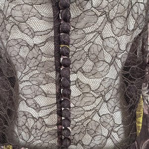 May include: A dark brown dress with a floral pattern and a sheer black lace overlay. The dress features a row of small, dark buttons down the front and a high neckline. The lace detailing adds a delicate touch to the garment.