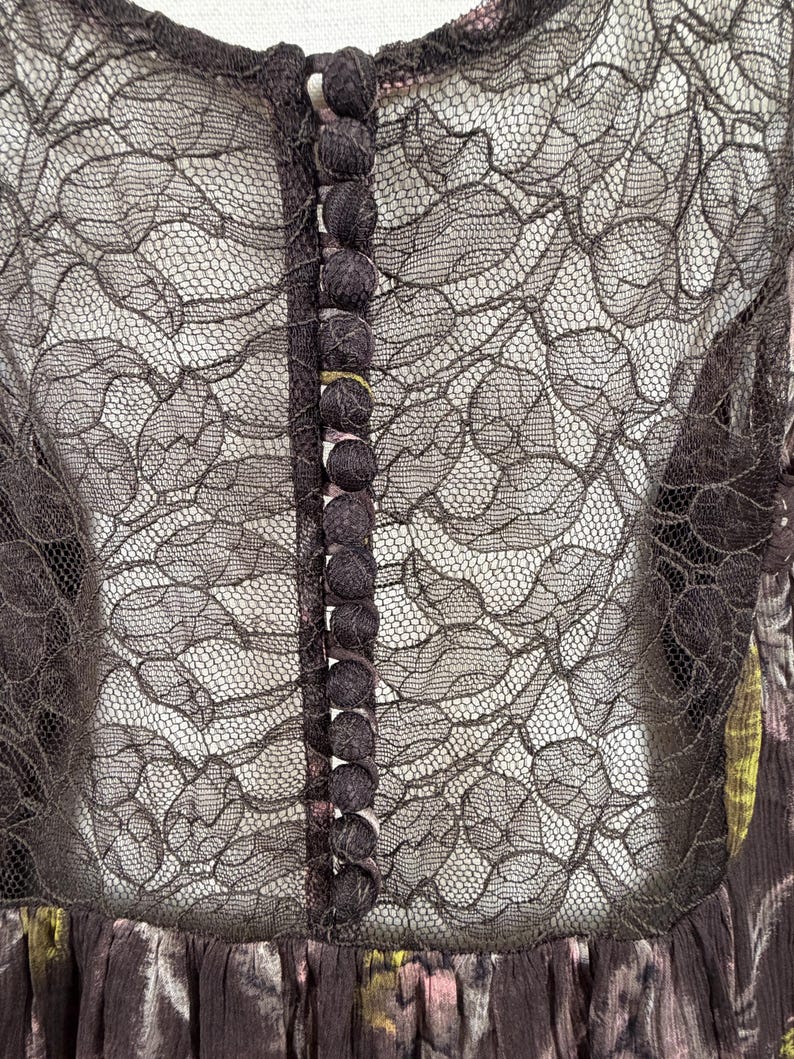 May include: A dark brown dress with a floral pattern and a sheer black lace overlay. The dress features a row of small, dark buttons down the front and a high neckline. The lace detailing adds a delicate touch to the garment.