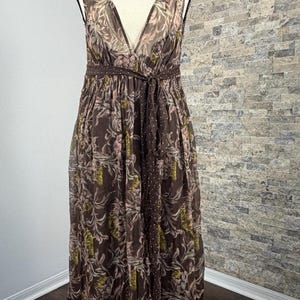 May include: A brown, sleeveless dress with a V-neck and a tie belt. The dress features a floral pattern with pink and green accents. The hemline has a polka dot design. The dress is displayed on a mannequin.