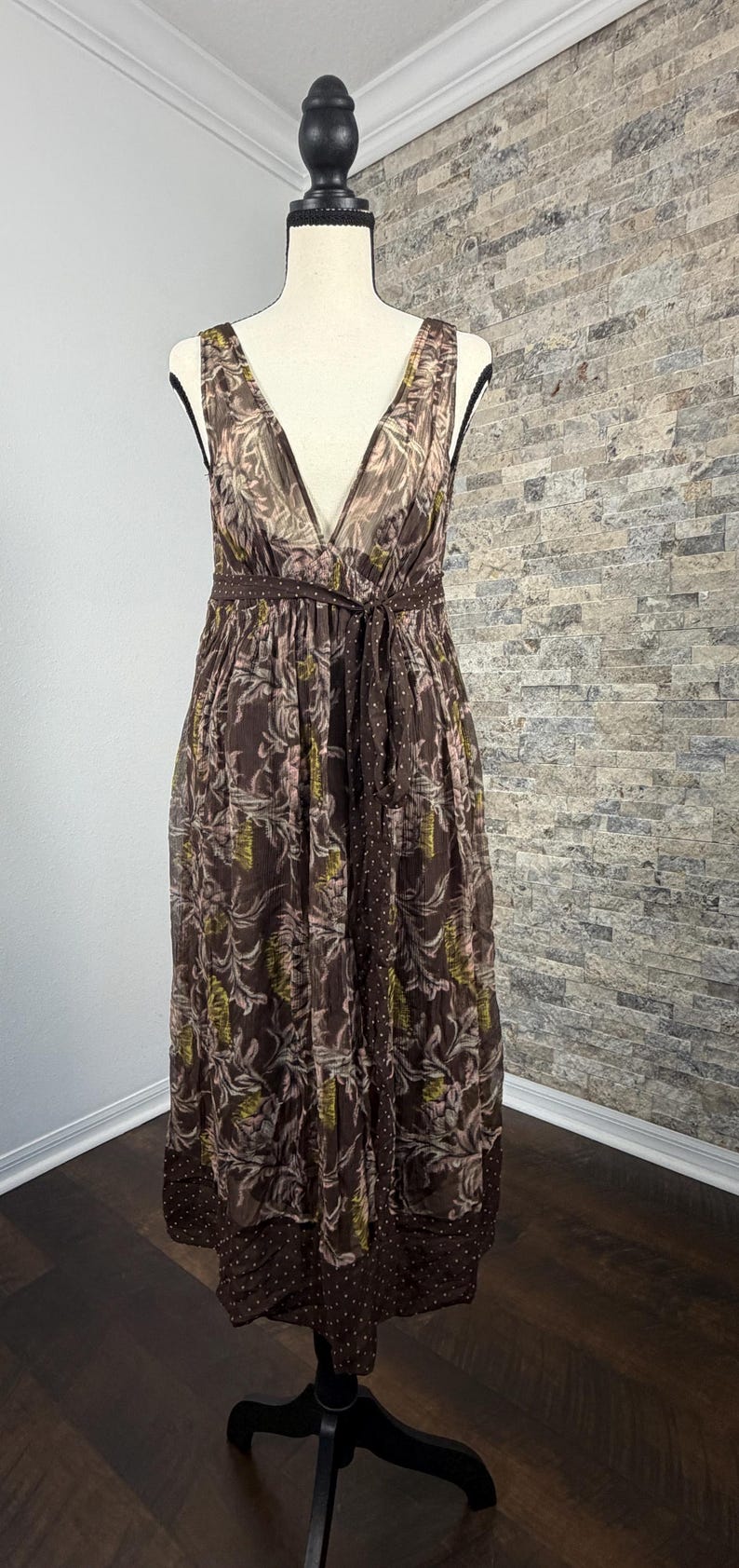 May include: A brown, sleeveless dress with a V-neck and a tie belt. The dress features a floral pattern with pink and green accents. The hemline has a polka dot design. The dress is displayed on a mannequin.