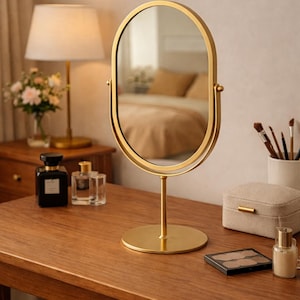 May include: A gold-coloured oval vanity mirror with a round base and stand. The mirror sits on a wooden table, alongside a lamp, perfume bottles, and makeup accessories. The room has a neutral colour palette.
