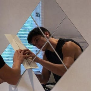 May include: A person is installing a set of diamond-shaped mirrors on a white wall. The mirrors reflect the person and the surrounding environment, including a window with blinds. The person is wearing a black tank top.