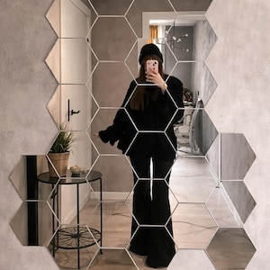 Puede incluir: Un espejo de pared moderno compuesto por baldosas hexagonales. El espejo está montado en una pared gris. Una pequeña mesa negra con tapa de cristal y una planta en maceta son visibles en el reflejo.