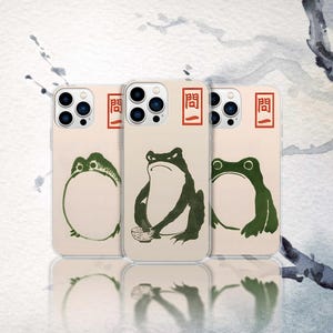 May include: Three phone cases with a beige background and green frog illustrations. Each case has a different frog design, with the text "問一" in a red rectangle at the top. The cases are arranged in a row.