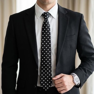 Peut inclure: Un costume noir avec une chemise blanche et une cravate noire à pois blancs. Une montre-bracelet argentée est visible. La veste de costume est boutonnée.